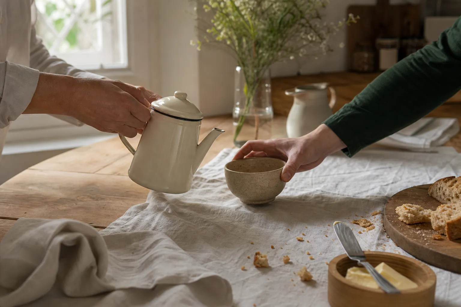 Deux mains posant une théière sur une table de cuisine en bois clair, lumière du matin par la fenêtre