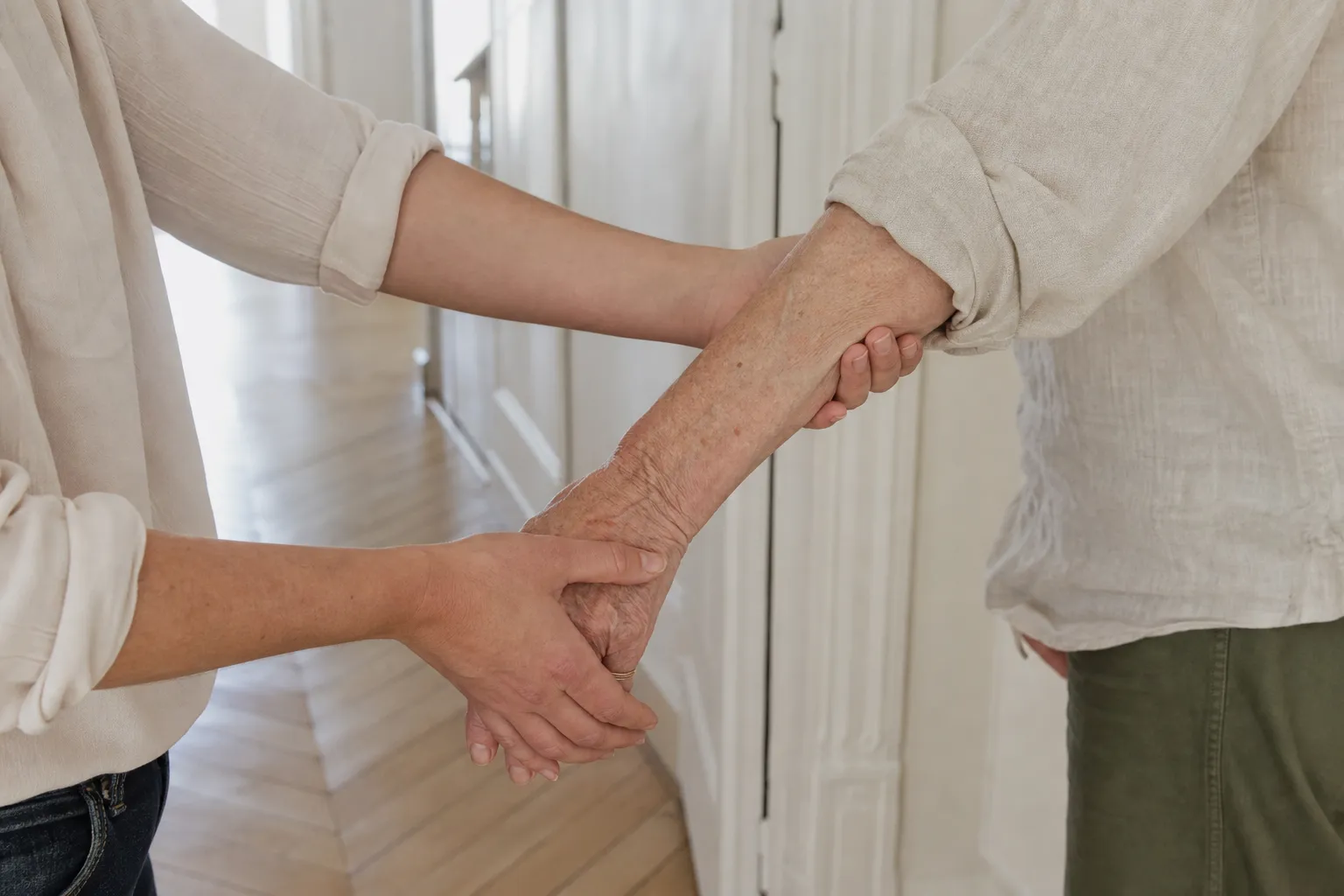 Une personne accompagnée par le bras dans le couloir clair d'un appartement, cadrage sur les mains et le pas